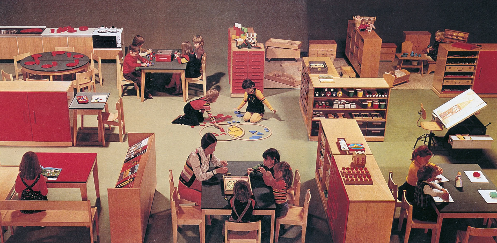 Kinder spielen in einem Kindergartenraum, der mit Raumteilern, Tischen und Stühlen ausgestattet ist.