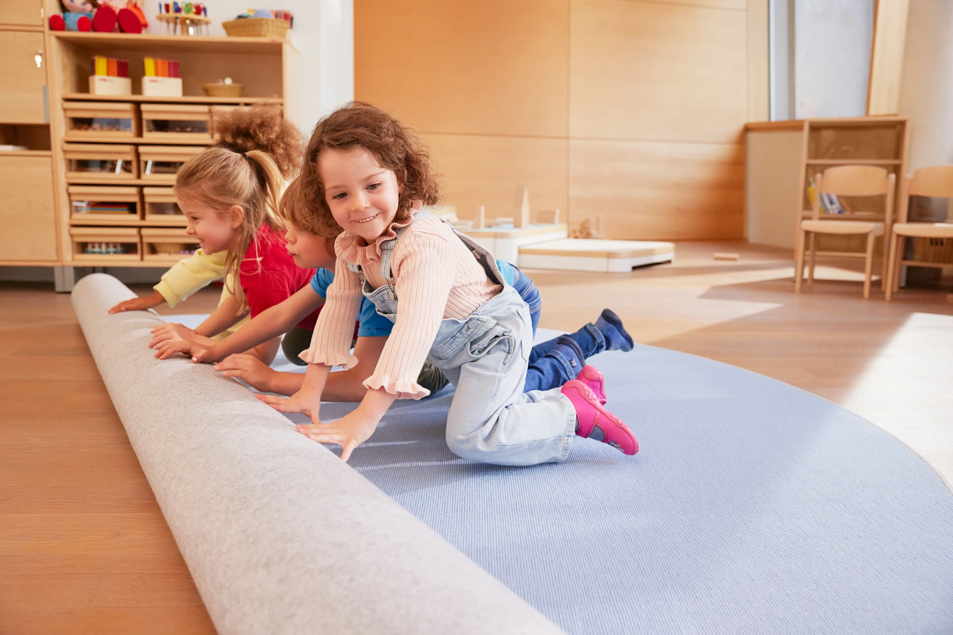 Kinder in einem Kindergartenraum rollen einen Teppich aus.