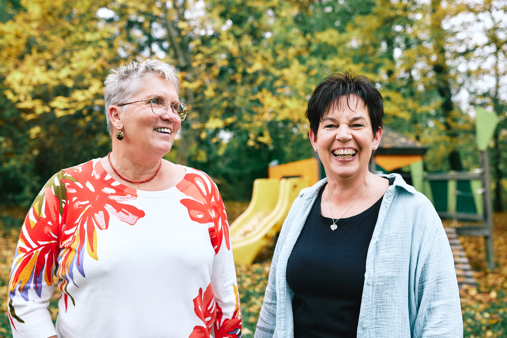 Beatrix Hirsch und Bianca Kupfer stehen lachend in einem Garten mit Bäumen und einer Rutsche.