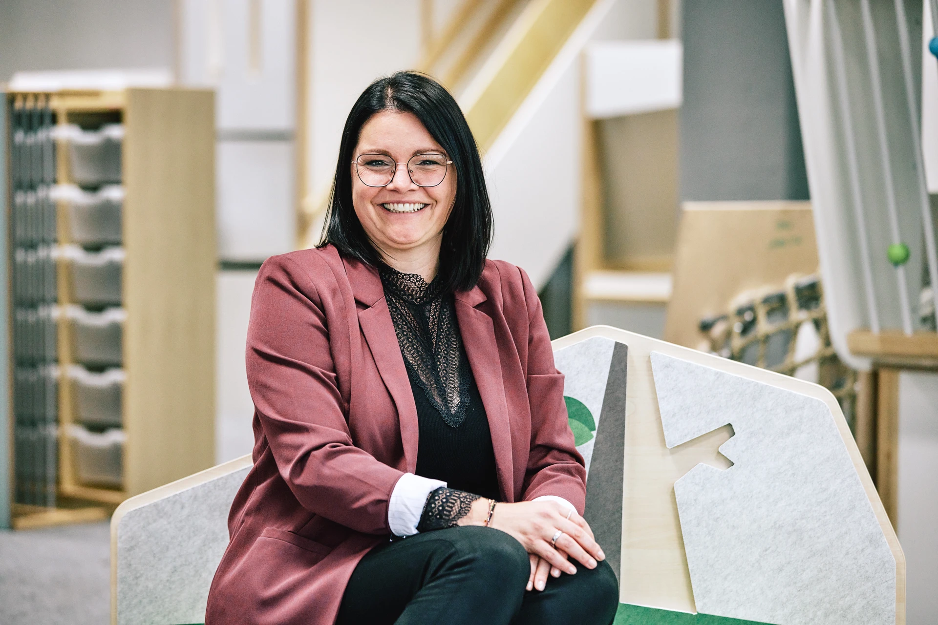 Janina Kopp sitzt auf einem Möbelstück der Firma Wehrfritz. Sie hat braune Haare, trägt eine Brille und einen roten Blazer.
