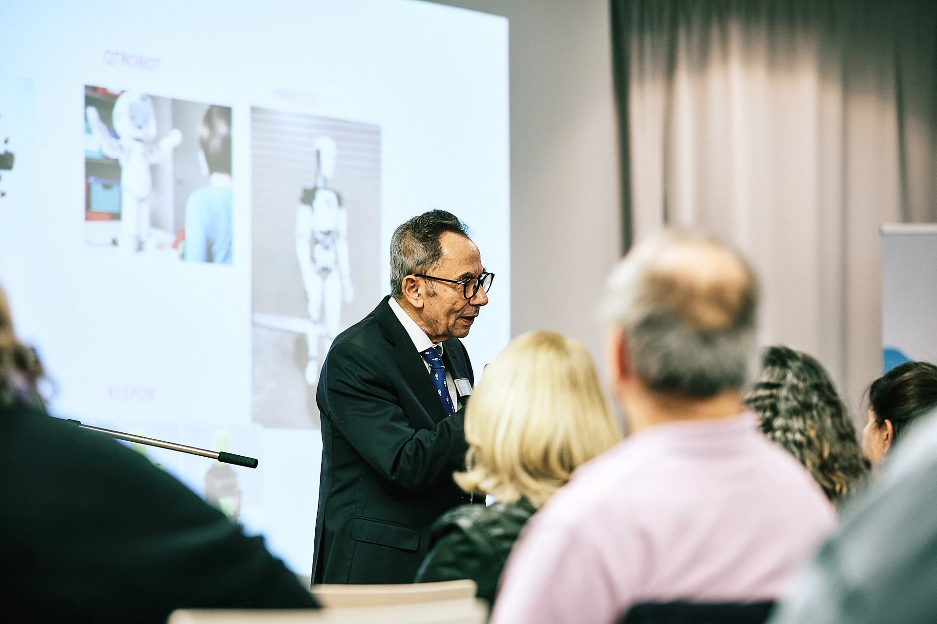 Der Bildungsforscher Prof. Dr. mult. Wassilios E. Fthenakis hält einen Vortrag vor Publikum. Hinter ihm sind auf die Wand Fotos von Robotern projiziert.